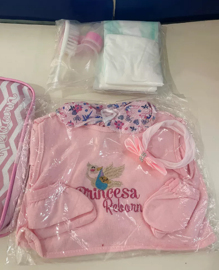 Itens do enxoval de bebê reborn com roupa rosa, fraldas e acessórios, mostrando o que compõe o valor da experiência de box opening.