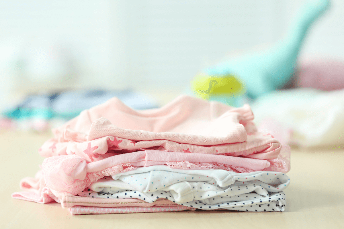 Roupas e enxoval que valorizam o kit bebê reborn Pilha de roupinhas de bebê dobradas em tons de rosa e branco, representando o enxoval e os acessórios que agregam valor ao preço final de um bebê reborn.