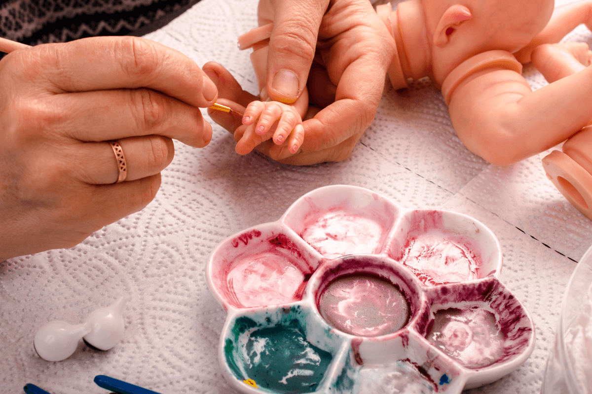 Mãos de uma artista pintando delicadamente as unhas de um kit, demonstrando o trabalho manual que define qual o valor original de um bebê reborn artesanal.