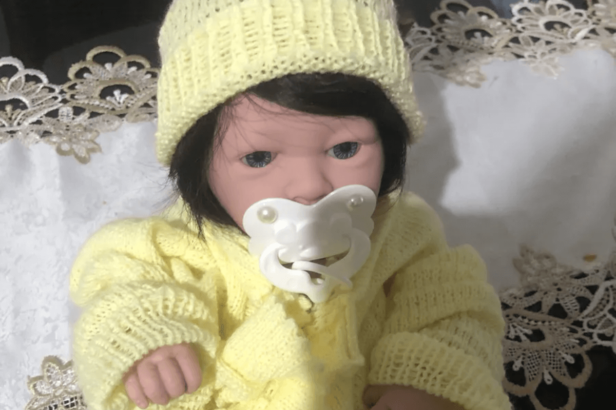 Guia sobre como escolher um bebê reborn, ilustrado por uma boneca realista com cabelo escuro e roupa de lã amarela, deitada em uma superfície com detalhes em renda.