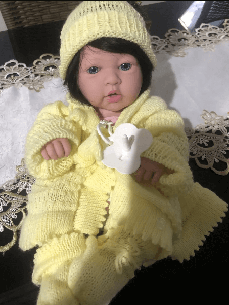 Bebê reborn com cabelo escuro implantado, possivelmente de mohair, vestindo um conjunto de lã amarela e touca combinando.