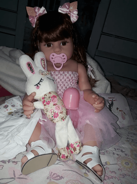 Foto caseira tirada por cliente mostrando uma boneca bebê reborn industrializada com coelho de pelúcia na cama, exemplificando o tipo de imagem real que deve ser exigida para comprovar que o vendedor possui o produto e evitar golpes com fotos roubadas.